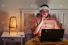 一个生病的成年男子在床头灯的灯光下对着屏幕做了一个可以接受的手势