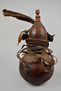 A Kwere medicine gourd from Tanzania. Here a calabash has been trained with a tight chord around its middle to take an hourglass shape so it can be suspended. The carved wooden stopper, depicting a mother with a baby on her back, identifies the gourd and 