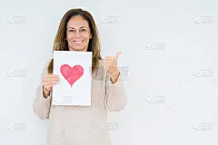 中年妇女手持卡片礼物与红心在孤立的背景指向和显示与拇指向上的一边与幸福的脸微笑