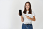 jovem-morena-mostrando-a-tela-do-smartphone-em-branco-e-apontando-o-dedo-para-o-celular-sorrindo-satisfeito-com-a-camera-em-pe-sobre-fundo-branco