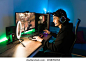 Smiling young man live streaming his online video game using a smartphone and a ring light. Happy gamer ready to start playing in a gaming computer