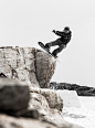 A snowboarder performs a trick and stalls on a boulder on a snowy mountain