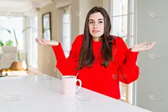 美丽的年轻女子喝着一杯黑咖啡，毫无头绪和困惑的表情与手臂和双手举起。怀疑的概念。