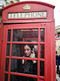 Woman in Black and White Floral Shirt Standing Inside Red Telephone Booth Making Call