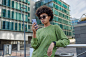 Stylish curly haired woman wears sunglasses and green jumper uses smartphone gets message poses against urban setting searches location via mobile phone application on street chats with followers