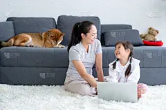 亚洲妇女和小女孩一起享受在客厅使用笔记本电脑和坐在地板上，而他们的柴狗躺在沙发上