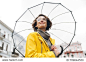View from beneath of positive woman in yellow raincoat and glasses standing in street under big transparent umbrella, during grey rainy day
