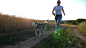 多莉拍摄了一个戴着墨镜的快乐女人和她的宠物沿着金色草地附近的道路奔跑。可爱的西伯利亚哈士奇狗在日落时分沿着麦田附近的道路慢跑时拉着皮带。慢动作