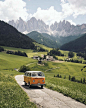 1️⃣ Volkswagen "T2" driving in the Dolomites, green grass and white flowers on both sides of it, with small villages scattered between them. The mountains rise high above the horizon, creating an epic view. A vintage orange van is parked at one 