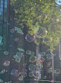 This may contain: soap bubbles are floating in the air near a brick building and green trees on a sunny day