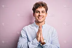 年轻的金发英俊的卷发男子穿着条纹衬衫在白色的背景祈祷与双手一起请求原谅自信的微笑。