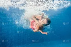 妈妈抱着女儿都浸在水里，在水中划桨。潜水的婴儿。幼儿学游泳。年轻的母亲或游泳教练和快乐的小女孩。