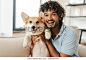 Young adult Indian man is smiling while holding his pet welsh pembroke corgi dog. They are both happy to be together at home. Love animal concept