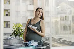 亲情母爱母爱，温情脉脉。母性本能，家务和孕妇。一位兴奋的准妈妈在家里的阳台上为新生儿准备衣服并摆姿势