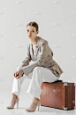 时尚女性旅行者在亚麻夹克坐在老式的手提箱在灰色背景隔离 