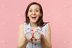 在画室里，一名穿着夏装、心情愉快的年轻女子，手里拿着半只新鲜成熟的火龙果，火龙果单独放在粉彩背景上。