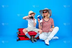 长胖快乐的女人和男孩，穿着夏天的衣服和帽子，护照和机票坐在红色的手提箱。家庭旅游和旅游。隔离在蓝色背