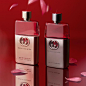 A pair of rose silver and chrome colored bottles of Gucci Guilty fragrance are arranged neatly on a diagonal upon a red mirrored surface where they are reflected in front of a matching red background, while pink rose petals fall from above. The flask-shap