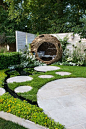 woven willow bird hide (willow sculpture) and concrete circular slabs as a path over a pond surrounded by Chamaemelum nobile (chamomile lawn), Eryngium giganteum, Eremurus himalaicus