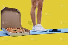 肌肉健美的女人的腿站在瑜伽垫上，旁边是一盒美味的新鲜披萨和哑铃。减肥和变胖的概念。健身与饮食概念。