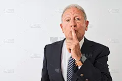 身穿西装的资深银发商人站在孤立的白色背景下，手指放在嘴唇上要求安静。沉默与秘密的概念。