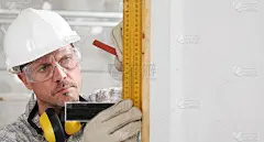 建筑工人用木工方尺和铅笔测量，戴安全帽、眼镜和听力保护耳机，在室内建筑工地