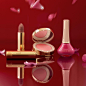 Several Gucci Beauty items are arranged playfully on a red mirrored surface where they are reflected in front of a matching red background, while pink rose petals fall from above. A gold tube of matte milk chocolate toned lipstick stands behind a striated