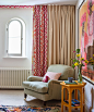 Girl's bedroom with pink patterned and cream curtains, small arched window, beige cozy armchair, cream carpet, colorful wall art, bright yellow side table