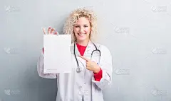 年轻的金发医生女人越过肮脏的灰色墙壁拿着空白的纸很高兴的指着手和手指