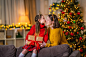 loving daughter kisses mom at home, sitting on couch against background of a Christmas tree. child and woman laughing happily, holding gift box in their hands Give each other gifts for Christmas