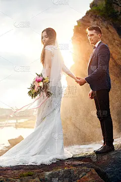一对美丽的情侣站在海边的岩石上接吻。新婚夫妇在夕阳和海浪下，爱和柔情。爱的夫妻休息。在户外婚礼。完美