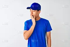 一名身穿蓝色t恤和帽子的中国快递员站在孤立的白色背景下无聊地打哈欠，疲惫地用手捂着嘴。不安和困倦。
