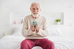 老人的照片坐在家里的床在室内快乐积极的微笑穿睡衣快乐积极的微笑使用浏览聊天类型短信智能手机