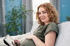 年轻孕妇使用平板电脑搜索怀孕信息。妈妈在家里的客厅沙发上躺着，一边照顾着孩子，一边开心地微笑，积极而