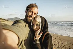 在日落时分，一对相爱的旅行情侣在一个荒凉的海滩自拍——女人与磁性的凝视和她的潮人男友在寻找相机自拍的