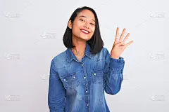 年轻美丽的中国女人穿着牛仔衬衫站在孤立的白色背景显示和指向第四个手指，同时自信和快乐的微笑。