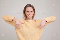 金发女人的肖像指向下微笑友好，凝视着相机建议尝试产品而不是看在灰色背景上的拷贝空间。穿黄色的毛衣。面