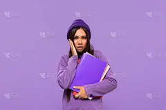漂亮的少年戴着帽子和毛衣，拿着文件夹，表达惊讶的情绪，站在紫色的背景孤立。教育的概念