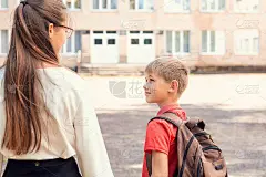 姐姐带弟弟去学校。男孩背着书包，牵着女孩的手，看着她，走向教学楼的入口。