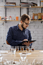 Portrait of man in glasses writing results of coffee cupping test, examining freshle ground coffee for flavour. He is standing near white wall in front of rows with glass cups