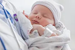 Sweet infant sleeps in doctor's armsAdorable Caucasian female infant is asleep in her doctor's arms. The baby is swaddled in a blanket and is wearing a cap. The unrecognizable doctor is wearing a lab coat.