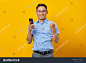Excited young handsome business man in classic blue shirt using mobile phone isolated over yellow background. Achieve career wealth business concept