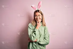 年轻美丽的女人戴着复活节兔子耳朵站在孤立的粉红色背景与手下巴思考问题，沉思的表情。带着沉思的微笑。怀