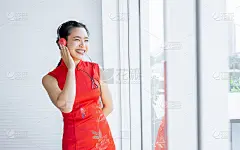 人像美丽开朗快乐的亚洲妇女穿着红色的服装，戴着耳机听音乐，面带微笑，独自站在室内的家庭或公寓的窗边，