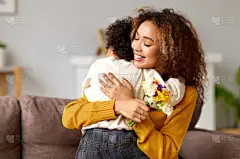 小儿子在家里和妈妈一起祝贺妈妈，送给她新鲜的花束，快乐的混血儿妈妈抱着儿子坐在沙发上。家庭假日和庆祝