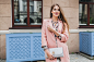 Cool young beautiful stylish woman walking in street in pink coat, holding purse in hands, listening to music
