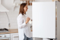 Free photo profile portrait of attractive dark haired woman wearing white shirt, looking smiling inside fridge with positive emotions, holding plate in hands, posing with kitchen set on background.