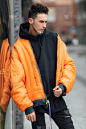时髦的男人站在建筑物附近的街道上。穿橙色夹克和全黑。冬天,秋天的衣服。夹克，衬衫和黑色运动鞋。