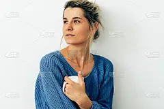 迷人的女人穿着舒适的针织蓝毛衣和一杯卡布奇诺的偷拍照片。漂亮的年轻女子在家里喝热饮。迷人的金发女人拿