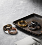 A color photo shows two David Yurman men’s band rings scattered on a grey marble surface next to a scratched, rusted metal dish holding three David Yurman men’s band rings. The jewelry is crafted from 18K yellow gold with or without forged carbon, 18K ros